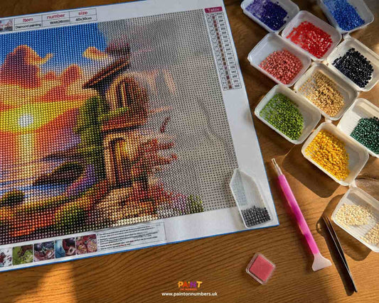 A diamond painting canvas in progress on a table, with trays of round resin diamonds sorted by colour and an applicator pen
