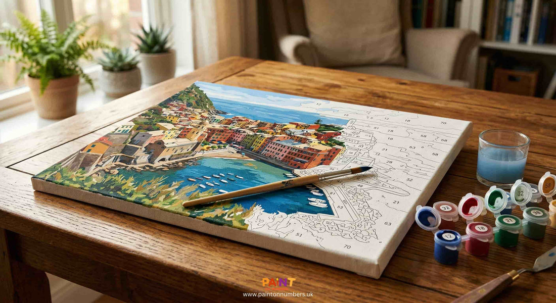 A partially completed paint by numbers canvas on a table showing several sessions of progress with brushes and paint pots beside it