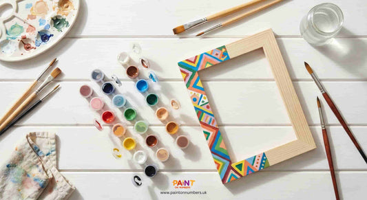 Paint by numbers DIY framing workspace with acrylic paint pots, artist brushes, painted wooden frame, and palette on a white wooden table.
