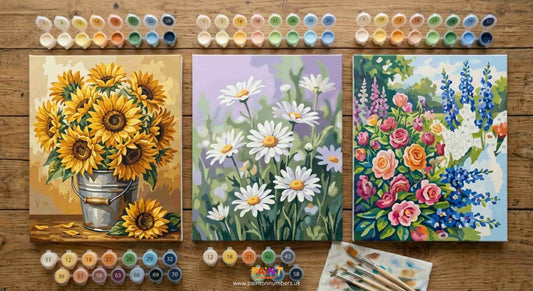 Selection of floral paint by numbers kits on a table showing completed canvases of sunflowers, daisies and botanical designs in various styles