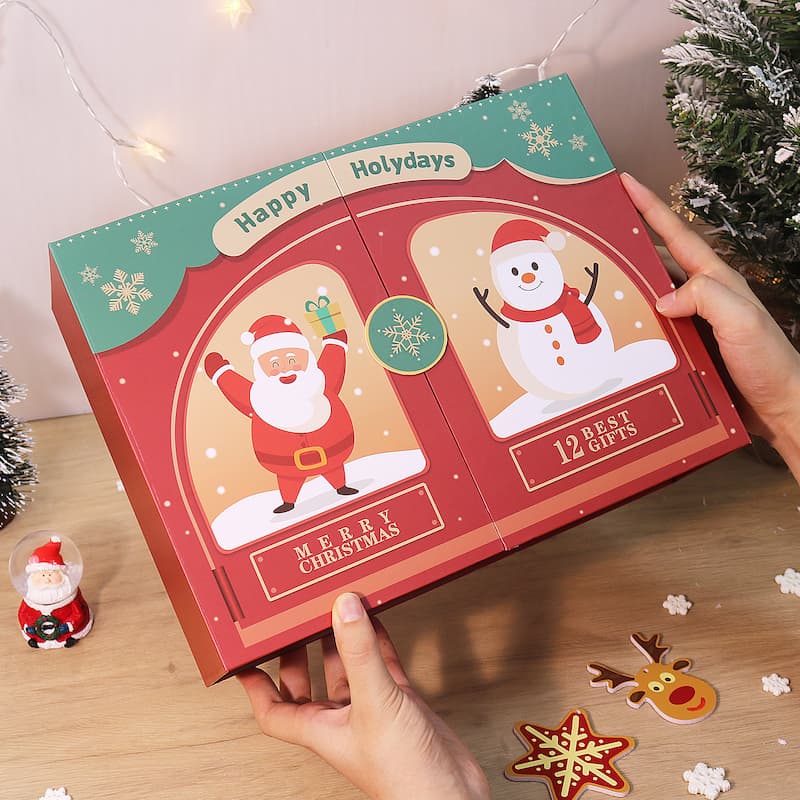 Person holding a festive red Christmas gift box featuring Santa and a snowman illustration with the text “Happy Holydays,” “Merry Christmas,” and “12 Best Gifts,” surrounded by holiday decorations on a wooden table.