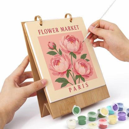 A hands-on painting activity setup featuring a bamboo easel with a canvas that reads 'FLOWER MARKET PARIS' and shows pink peony flowers. A person's hands are holding the easel and using a paintbrush, with small paint pots arranged on a white surface below.