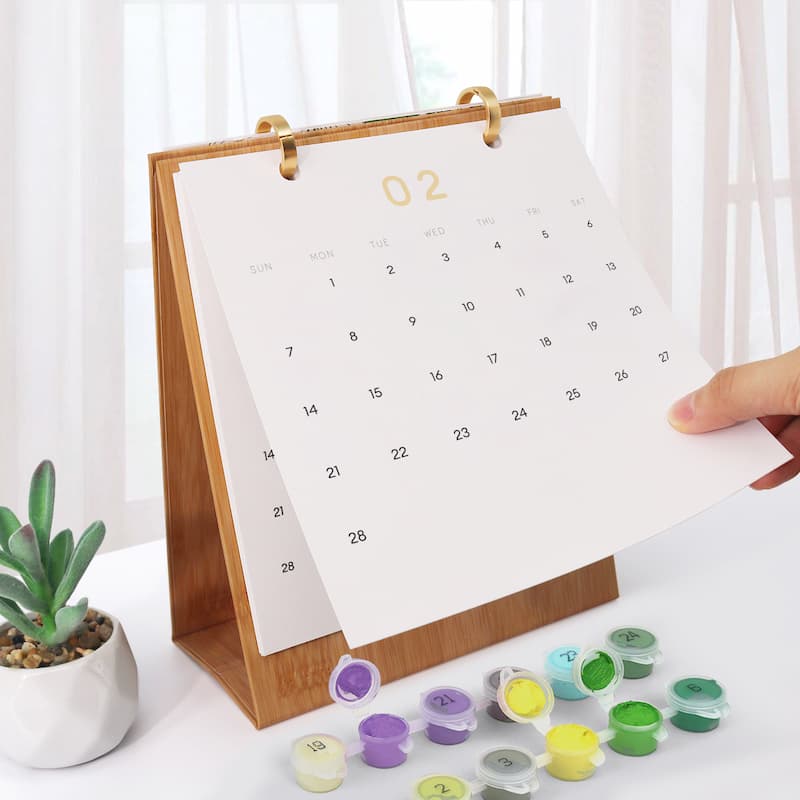 A wooden desktop calendar stand with a white page showing the number 2, held by a hand. Small paint pots in multiple colors are scattered in front on a white surface, with a small potted succulent and sheer curtains in the background.