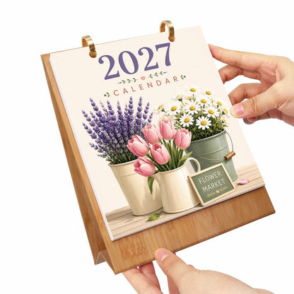 A 2027 desk calendar with a bamboo stand, held by two hands. The cover features an illustration of lavender, pink tulips, and white daisies in metal pots, with a small chalkboard sign reading 'FLOWER MARKET'.