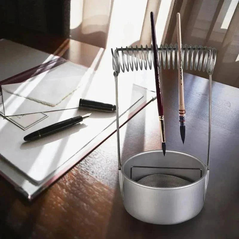 Metal pen holder with brushes on a desk next to a notebook and pen