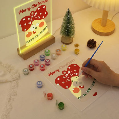 A hand is painting a Christmas-themed acrylic panel with a cartoon character and 'Merry Christmas' text, while a glowing version of the same panel stands beside it on a tabletop. Paint pots, a small tree, and a lamp are arranged around it.