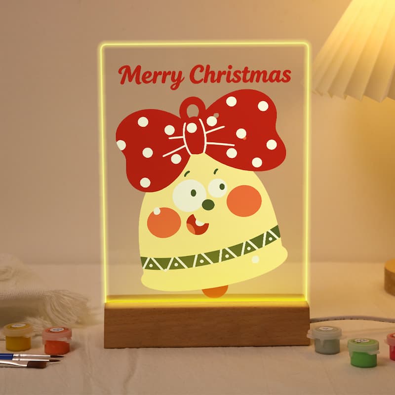 A glowing acrylic Christmas bell lamp on a wooden base, featuring a cartoon bell character with a red polka-dot bow and the words 'Merry Christmas' at the top, shown lit in a warm indoor setting.
