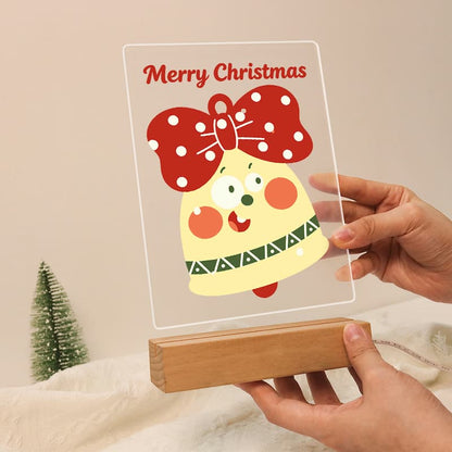 A clear acrylic LED light box displaying a festive Christmas design with a smiling yellow bell and red bow, resting on a wooden base held by two hands against a warm beige background.