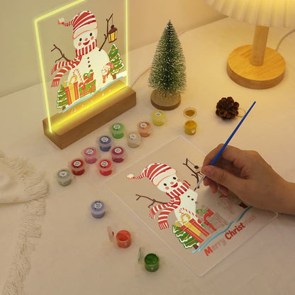A hand is painting a snowman design on a clear panel with a blue brush, placed on a table next to a lit acrylic LED snowman lamp, small Christmas trees, paint pots, and a lamp.