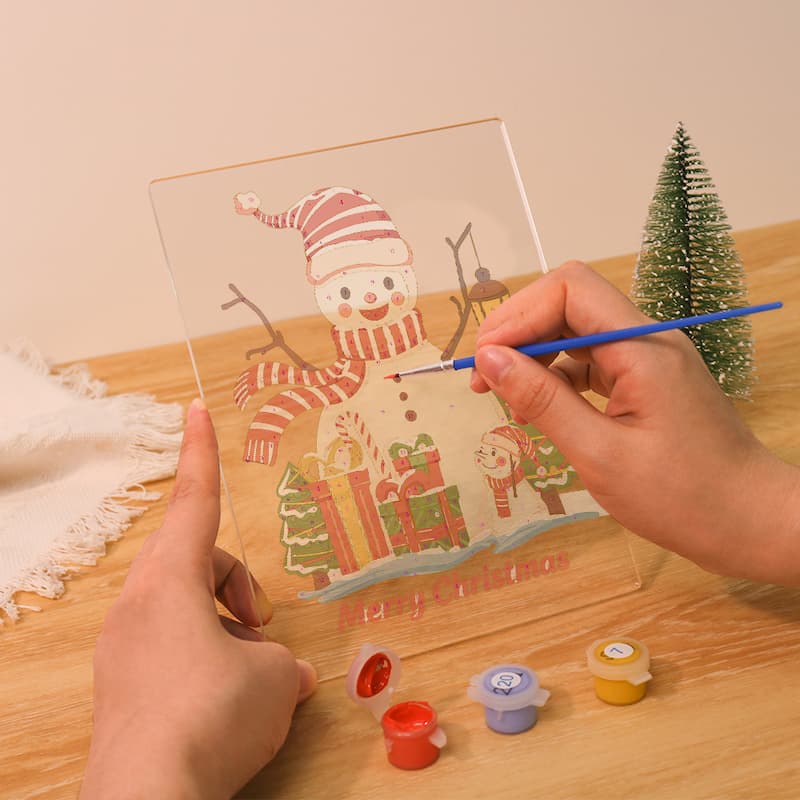 A person holds a clear acrylic canvas with a painted snowman design while using a blue paintbrush to add details. Small paint pots and a miniature Christmas tree sit on a wooden table in a warm indoor setting.