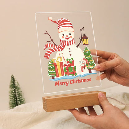 A clear acrylic Christmas sign featuring a smiling snowman with a red-and-white striped hat and scarf, wrapped in holiday greenery, and the words 'Merry Christmas,' displayed on a wooden block stand with hands holding it.