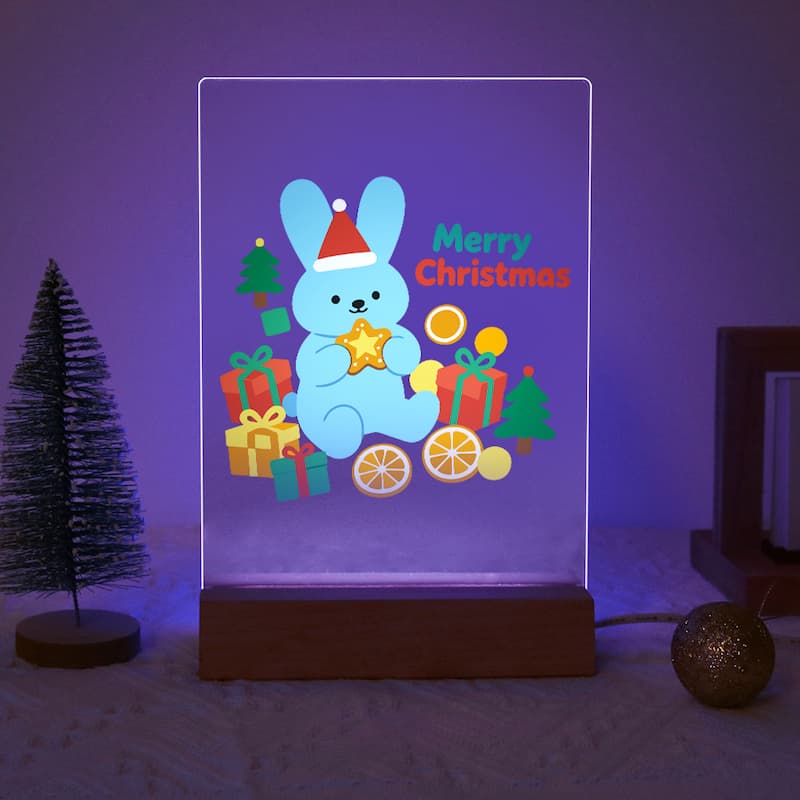 A rectangular LED light box on a wooden base displaying a cute light-blue bunny holding a gold star, surrounded by Christmas presents, trees, and the words 'Merry Christmas' in colorful text. The scene is set on a bed with a small tree and a gold ornament in a bedroom.