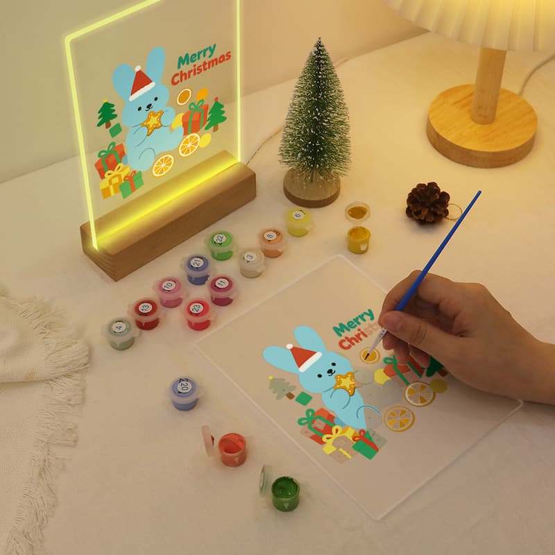 A hand is painting a Christmas-themed design on a clear acrylic panel with a blue-handled brush, while a matching illuminated acrylic sign stands nearby on a wooden base. The scene includes small paint pots, a mini tree, and a lamp on a tabletop.