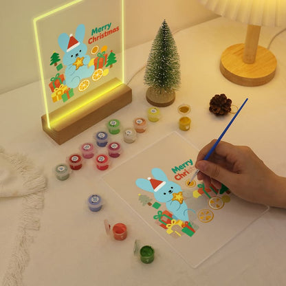 A hand is painting a Christmas-themed design on a clear acrylic panel with a blue-handled brush, while a matching illuminated acrylic sign stands nearby on a wooden base. The scene includes small paint pots, a mini tree, and a lamp on a tabletop.