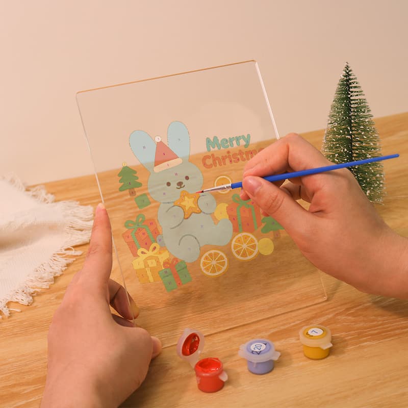 A person holds a clear acrylic painting plate with a cartoon bunny and 'Merry Christmas' design, using a blue paintbrush. Small paint pots and a miniature tree sit on a wooden table against a warm beige background.