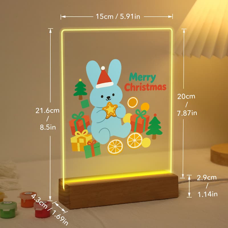 A glowing acrylic Christmas sign with a cartoon blue bunny holding a star, surrounded by presents, trees, and the words 'Merry Christmas,' mounted on a wooden base. White dimension lines and text show the product's height, width, and base depth in centimeters and inches.