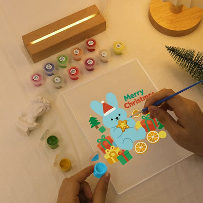 A person's hands are shown painting a light blue bunny with a Santa hat and a star, next to colorful Christmas-themed gift boxes and presents on a clear acrylic canvas. The scene is set on a light wooden table with various paint pots, a wooden light box, and a small green tree.