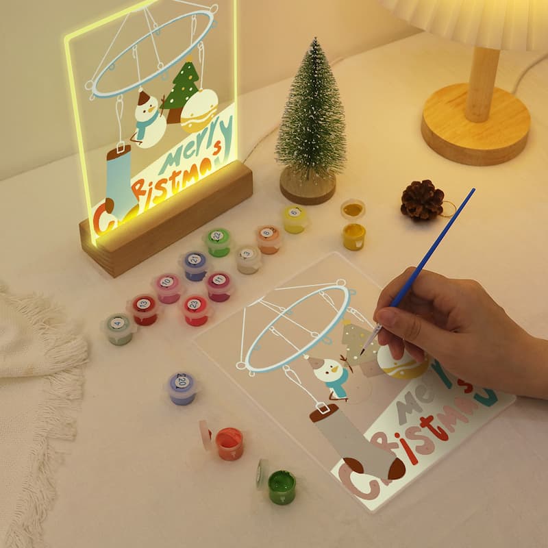 A hand is painting a clear acrylic panel with a Christmas scene, shown next to a completed, glowing LED light sign on a wooden base. The setup includes small paint pots, a miniature Christmas tree, and a lamp on a warm, neutral tabletop.
