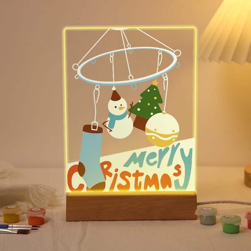 A wooden LED night light with a Christmas-themed design, including a snowman, Christmas tree, ornaments, and 'Merry Christmas' text, glowing warmly on a tabletop.