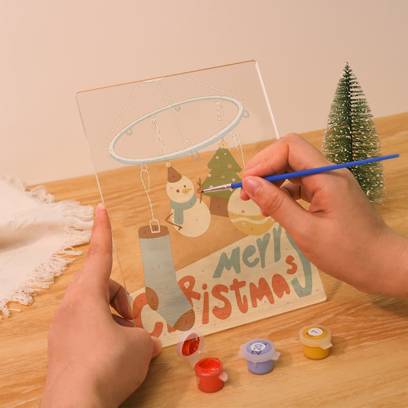 A person holds a clear acrylic Christmas ornament painting board with a painted snowman, tree, and 'Merry Christmas' design, while using a blue paintbrush. Small paint pots are placed below on a wooden table, with a small green tree prop and a cream background.