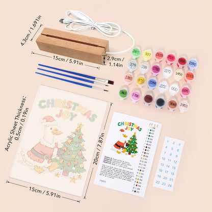 Christmas DIY canvas painting kit with a wooden frame, acrylic paint bottles, two paintbrushes, and a canvas sheet showing a cartoon duck decorating a Christmas tree, laid out on a beige background with printed dimensions.