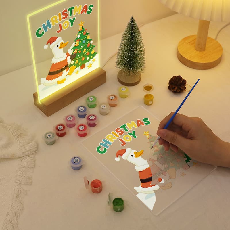 A Christmas-themed DIY acrylic painting kit shown on a tabletop, with a hand holding a blue brush over a clear panel featuring a cartoon duck and the words “Christmas Joy.” Several small paint pots, a small decorative tree, and a glowing framed artwork are arranged around it.