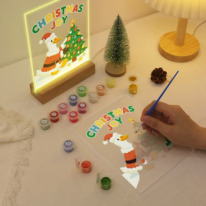 A Christmas-themed DIY acrylic painting kit shown on a tabletop, with a hand holding a blue brush over a clear panel featuring a cartoon duck and the words “Christmas Joy.” Several small paint pots, a small decorative tree, and a glowing framed artwork are arranged around it.