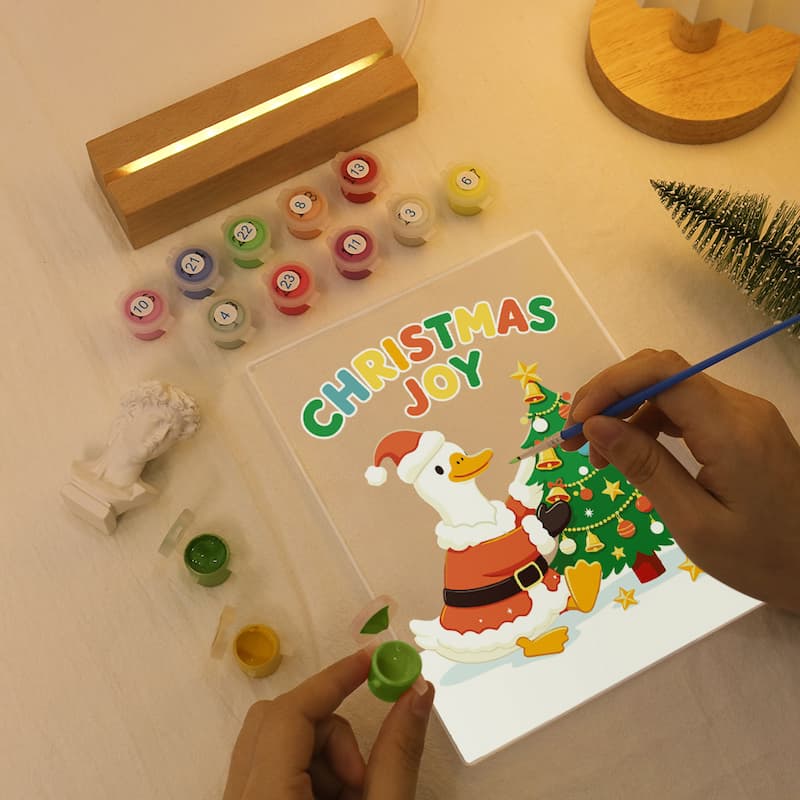 A Christmas-themed acrylic painting kit being used by two hands on a light-colored wooden table. The clear panel features a colorful illustration of Santa Claus and a Christmas tree with the words 'CHRISTMAS JOY' in multicolored letters.