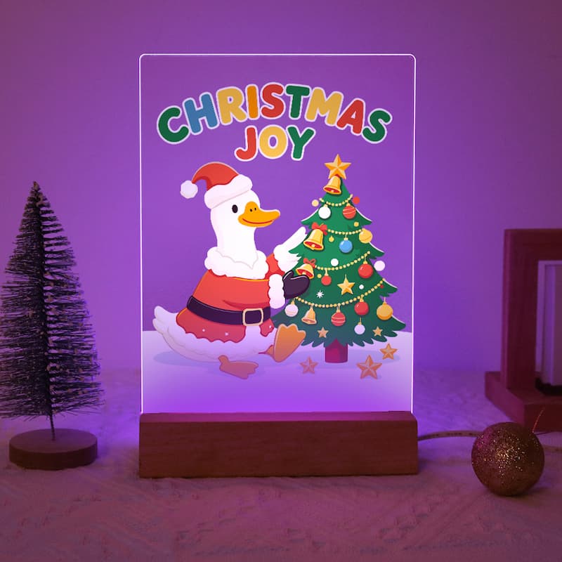A rectangular LED night light with a wooden base displays a colorful Christmas illustration featuring the text 'CHRISTMAS JOY' and a duck in a Santa outfit next to a decorated tree. The product is shown lit up in a purple glow on a textured surface with holiday props.