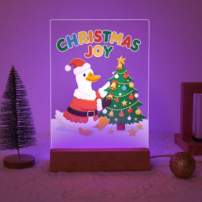 A rectangular LED night light with a wooden base displays a colorful Christmas illustration featuring the text 'CHRISTMAS JOY' and a duck in a Santa outfit next to a decorated tree. The product is shown lit up in a purple glow on a textured surface with holiday props.