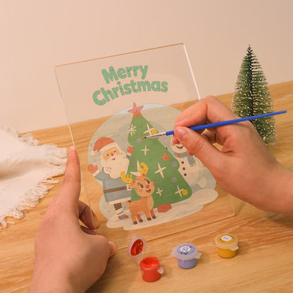A person holds a clear acrylic panel with a printed Christmas scene and the words “Merry Christmas,” while painting over the illustration with a blue-handled brush. Small paint pots and a miniature tree sit on a wooden table in a warm indoor setting.