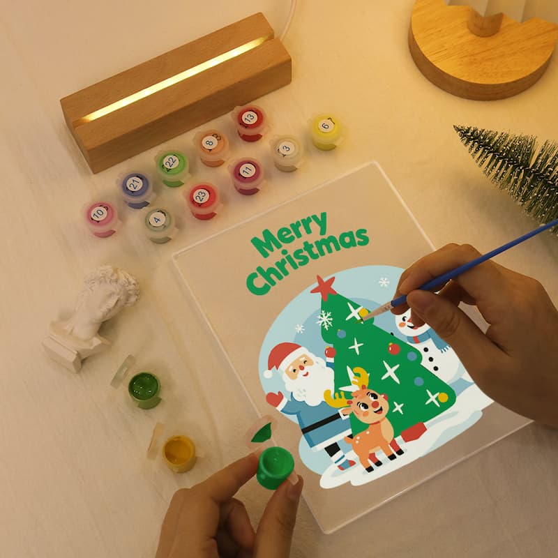 A person's hands are painting a Christmas-themed canvas print on a clear acrylic panel. The scene is styled as a flat-lay with a wooden light box, miniature Santa, colorful paint pots, and a small Christmas tree on a light wood surface.