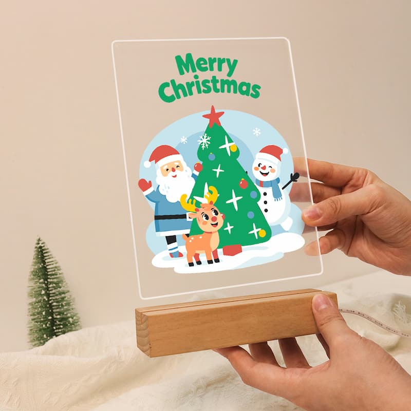 A clear acrylic Christmas sign on a wooden stand being held by hands. The sign features a colorful illustration of Santa Claus, a snowman, a reindeer, and a Christmas tree with the text 'Merry Christmas' at the top.