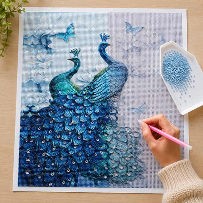 A person's hand holding a pink diamond painting pen works on a completed blue peacock canvas design, with a small dish of blue beads on a light wood tabletop.