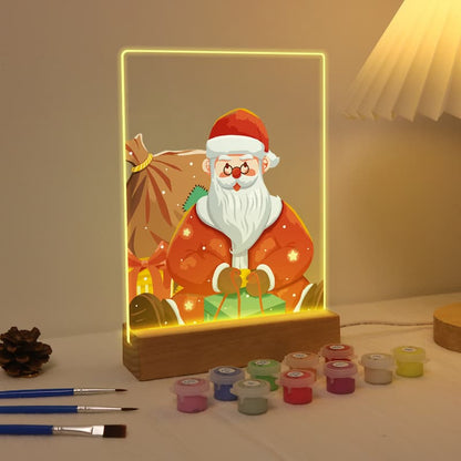 A 3D LED acrylic light box featuring a cartoon Santa Claus holding a gift, glowing with warm yellow light. The wooden base sits on a tabletop next to paint pots, paintbrushes, and a lamp in a cozy room setting.