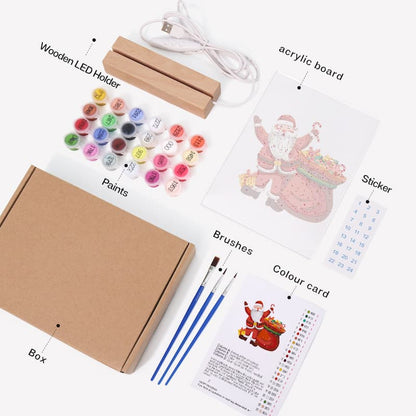 A Christmas-themed acrylic painting kit laid out on a white background, including a kraft paper box, wooden LED light holder, USB charging cable, acrylic board with Santa illustration, sticker, paint pots, brushes, color card, and instruction sheet.