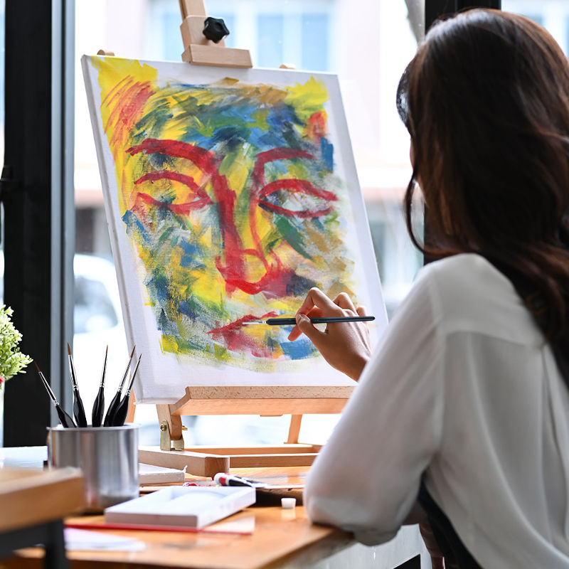 Artist at a wooden tabletop easel using a detail brush to paint on canvas with acrylic paint