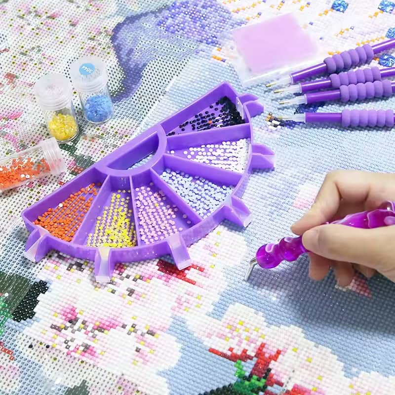 Half diamond painting sorting tray filled with colorful drills beside a diamond painting canvas while a crafter places drills using a diamond painting pen.