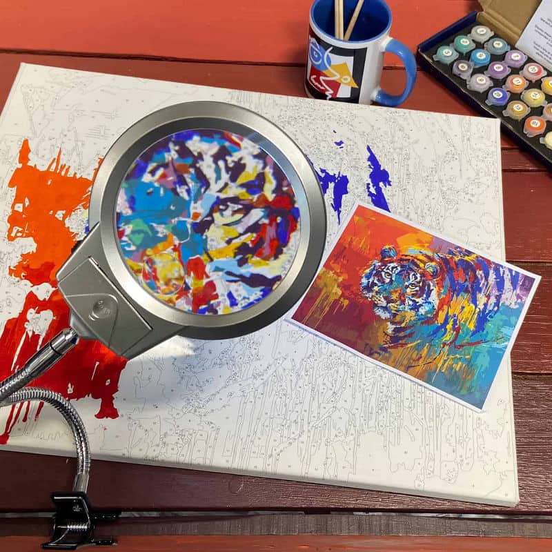 Magnifying glass over a colorful abstract painting on a wooden surface with art supplies.