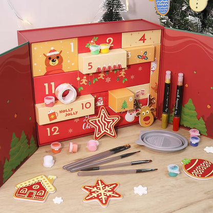 Christmas-themed advent calendar box with 12 numbered drawers, surrounded by paint brushes, mini paints, craft markers, and festive decorations like stars, reindeer, and gingerbread ornaments.